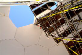 View from inside a radome with hexagonal panels, looking up at blue sky through an opening; scaffolding and caution tape at edge.