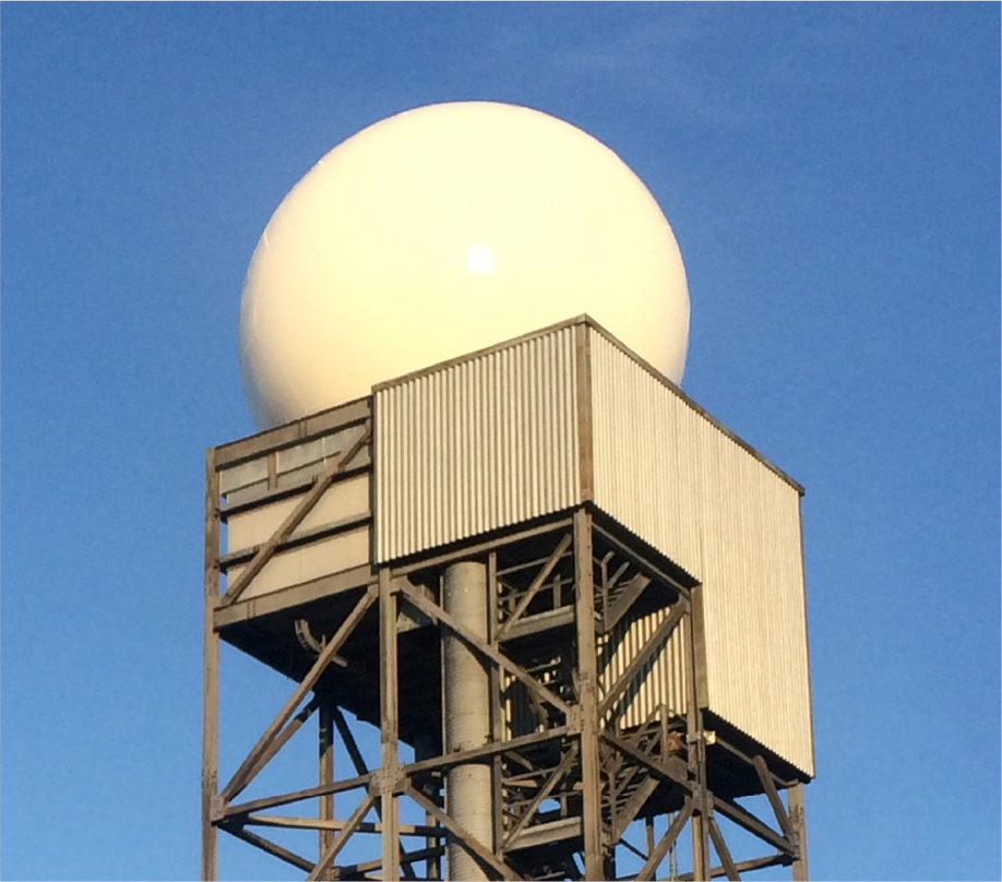A large white weather radar dome, used in various meteorological applications, sits atop a tall metal tower with corrugated metal siding, set against a clear blue sky.