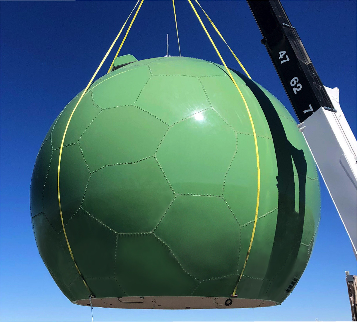 A large, green, dome-shaped structure with a pentagon and hexagon pattern is being lifted by a crane against a clear blue sky. Yellow straps are securing the dome as it is hoisted.